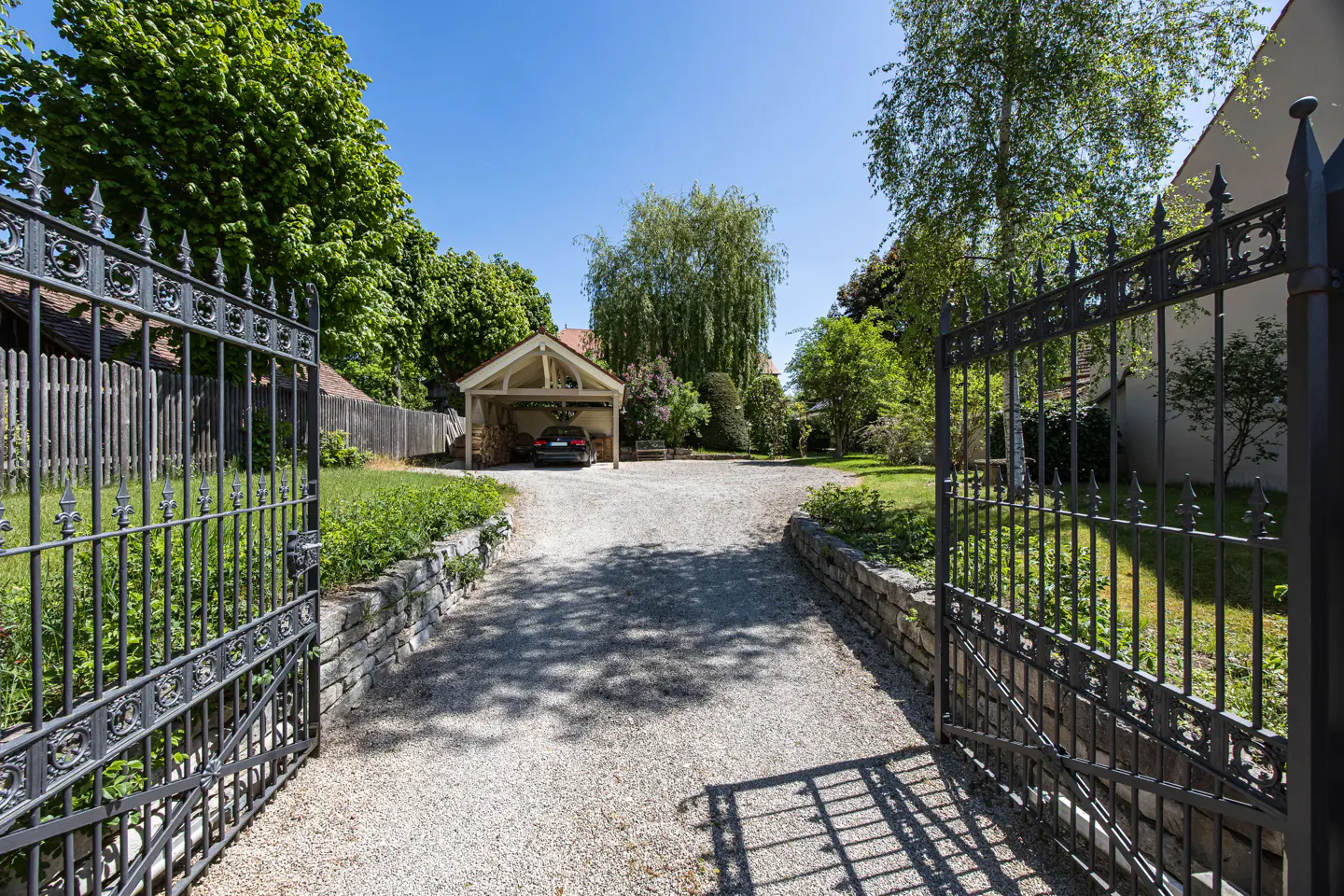 Open wrought iron gate leads to a gravel driveway, carport with car, and lush green trees under a blue sky.
