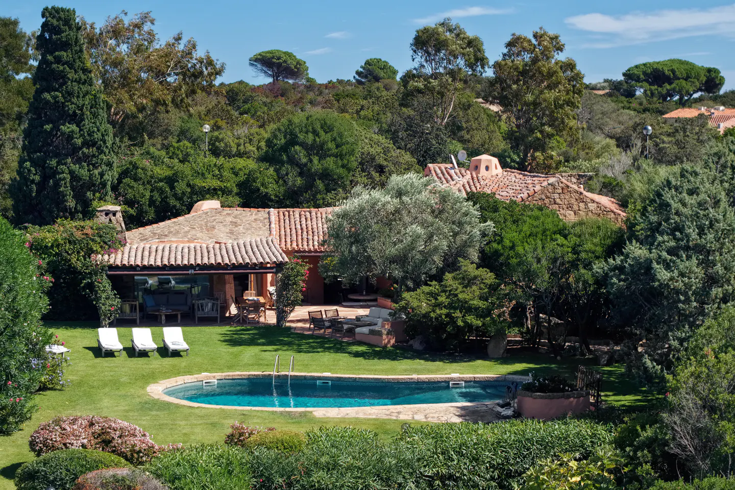A Mediterranean-style villa with a terracotta roof, a swimming pool, and lounge chairs on a lush green lawn.