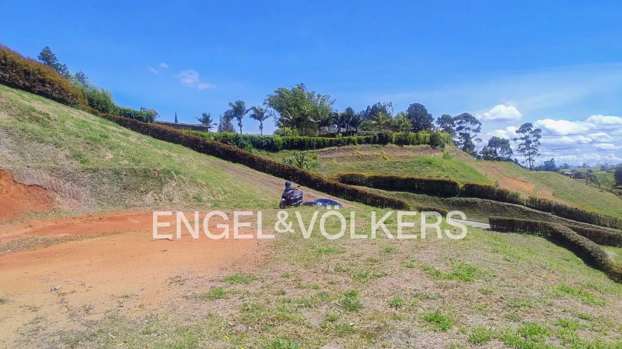 A grassy hillside with terraced hedges under a blue sky. A motorcycle is parked on the hill. The Engel & Volkers logo is superimposed.