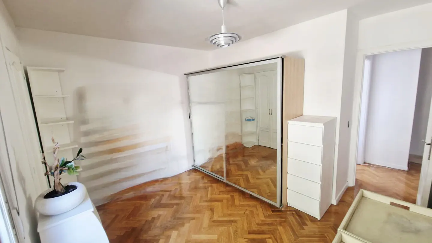 Bright room with herringbone wood floors, white walls, and a mirrored sliding door closet. A white dresser and shelves are also visible.