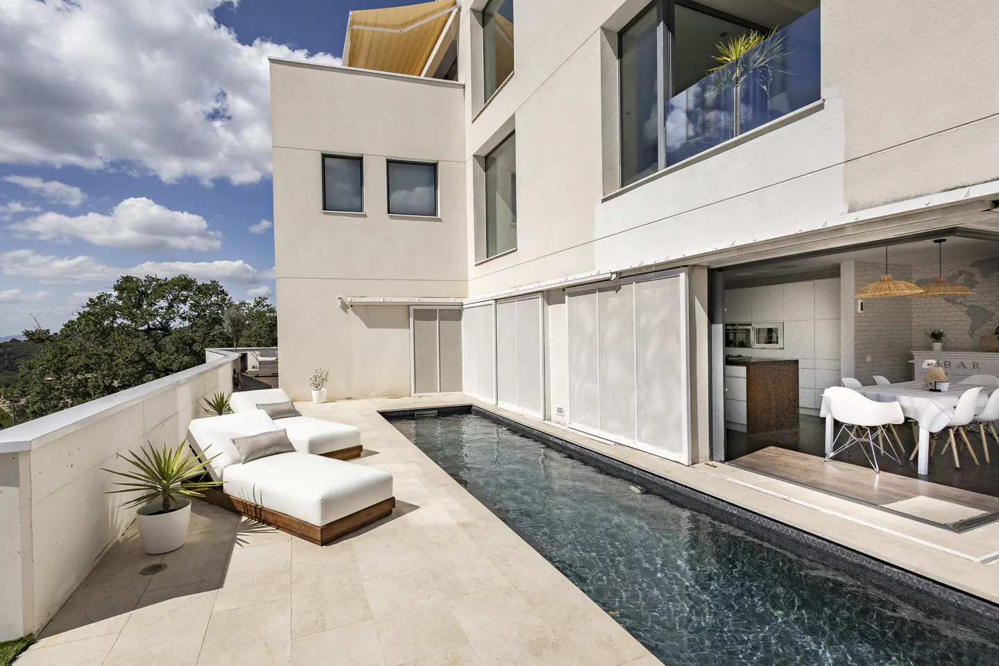 Modern white house with a narrow pool, lounge chairs, and an open doorway to a dining area.