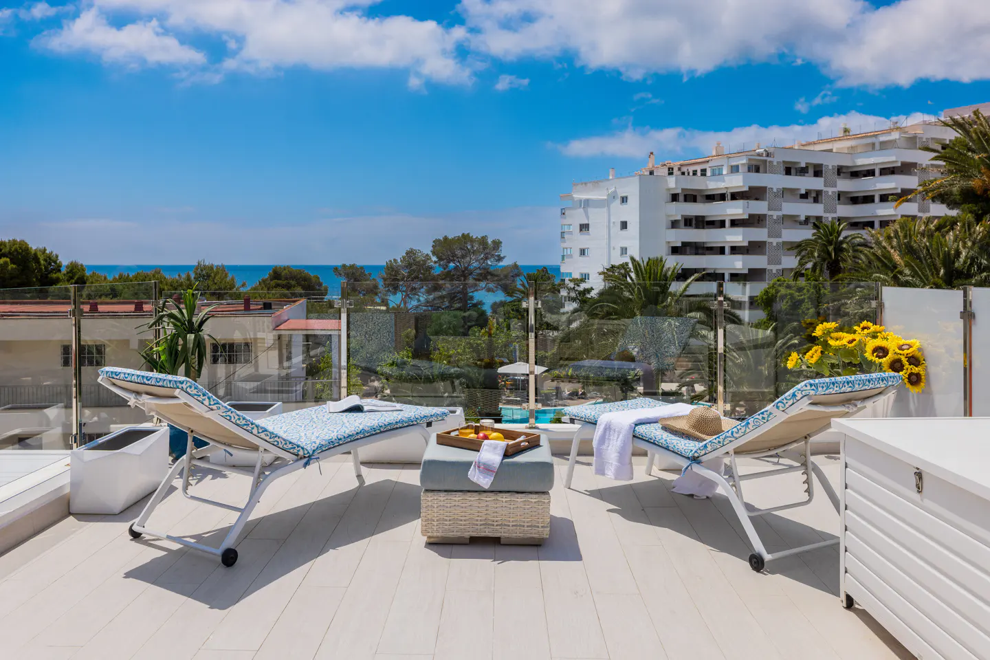 Rooftop patio with two blue-cushioned lounge chairs, a wicker ottoman with a fruit tray, and a view of the ocean and buildings.