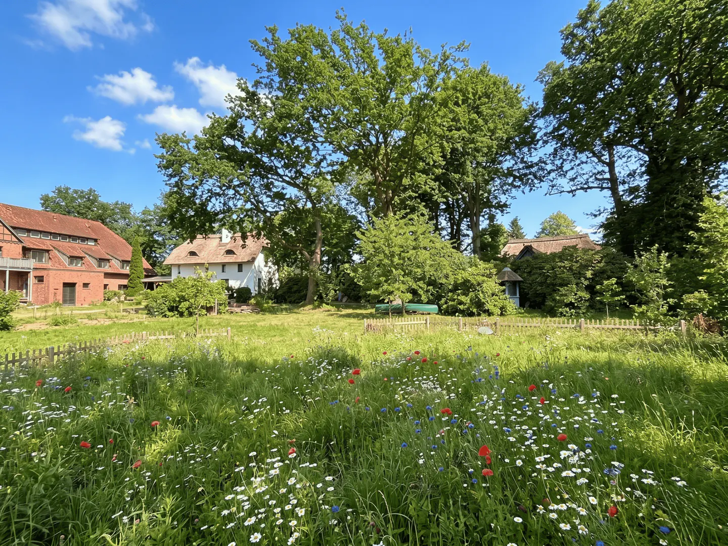 Lush meadow with wildflowers, a picket fence, and trees. In the background are a red brick building and a white house with a thatched roof.