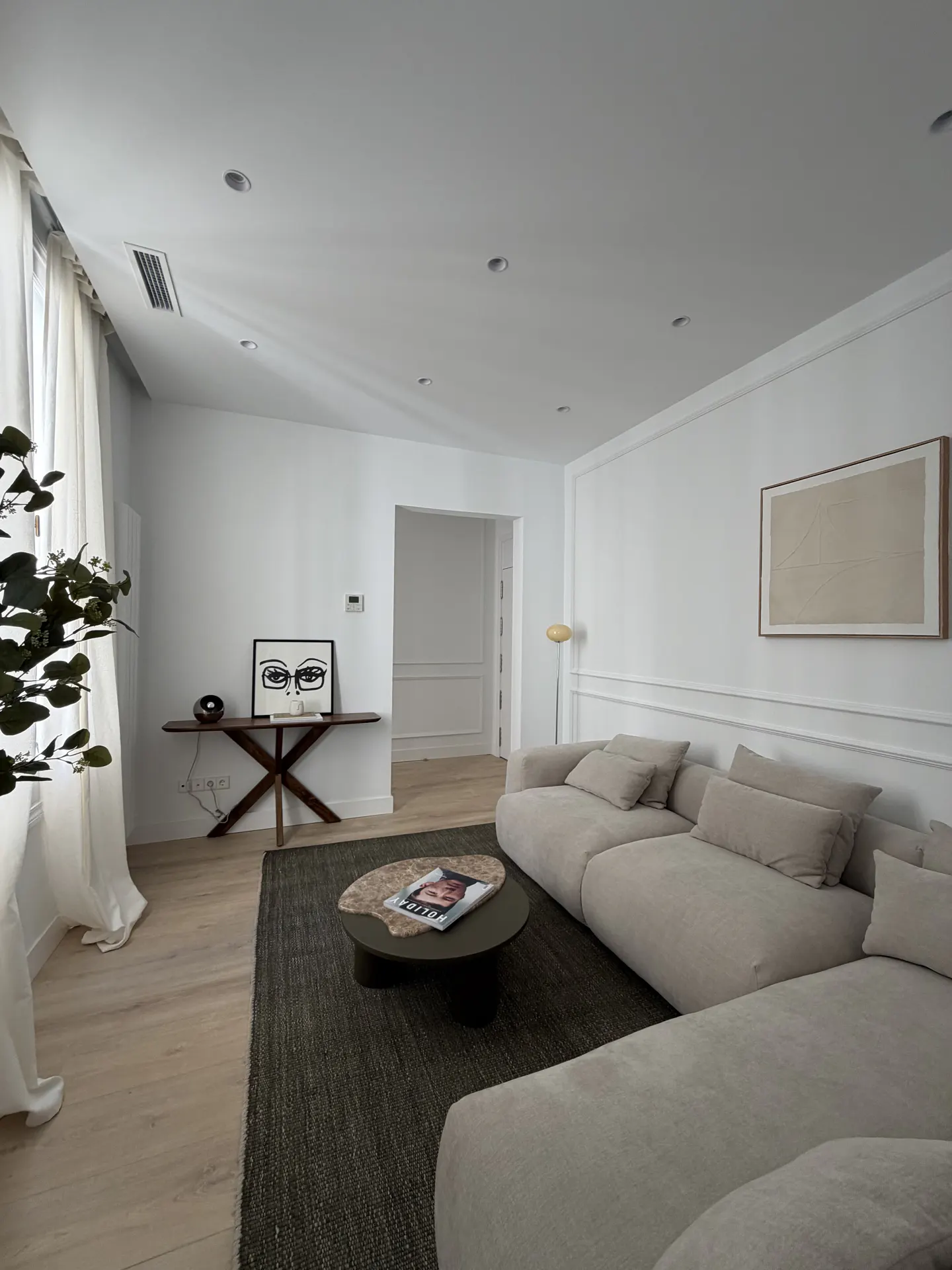 Bright living room with a large beige sectional sofa, dark rug, and a round coffee table. A console table with art sits against the white wall.