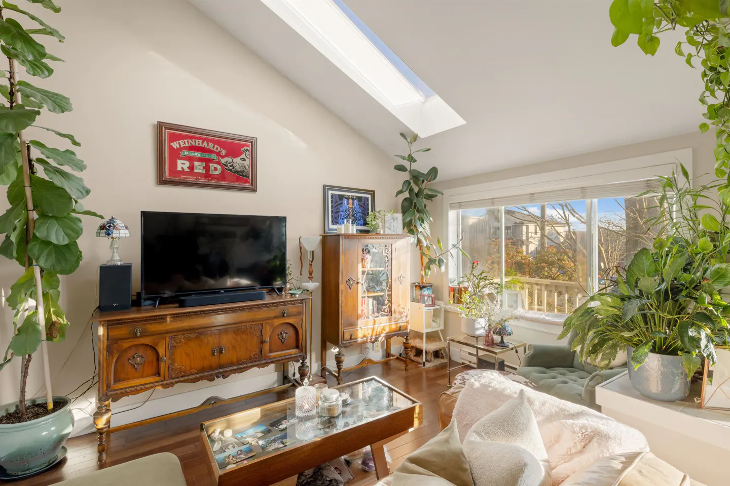 Bright living room with hardwood floors, a skylight, and many plants. A TV sits on a wooden console table.