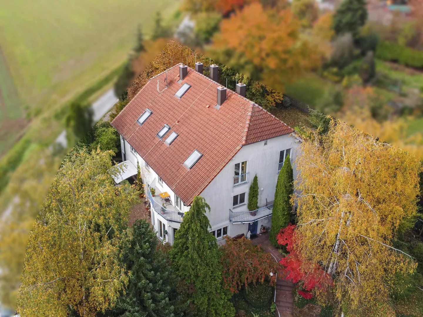 Aerial view of a white two-story house with a red tile roof, surrounded by colorful autumn trees.