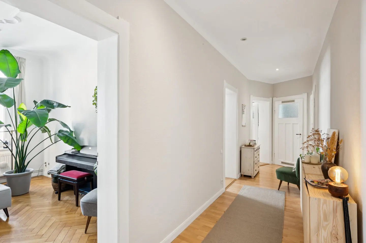 Hallway with light walls and wood floors. A living room with a piano and large plant is visible through a doorway.