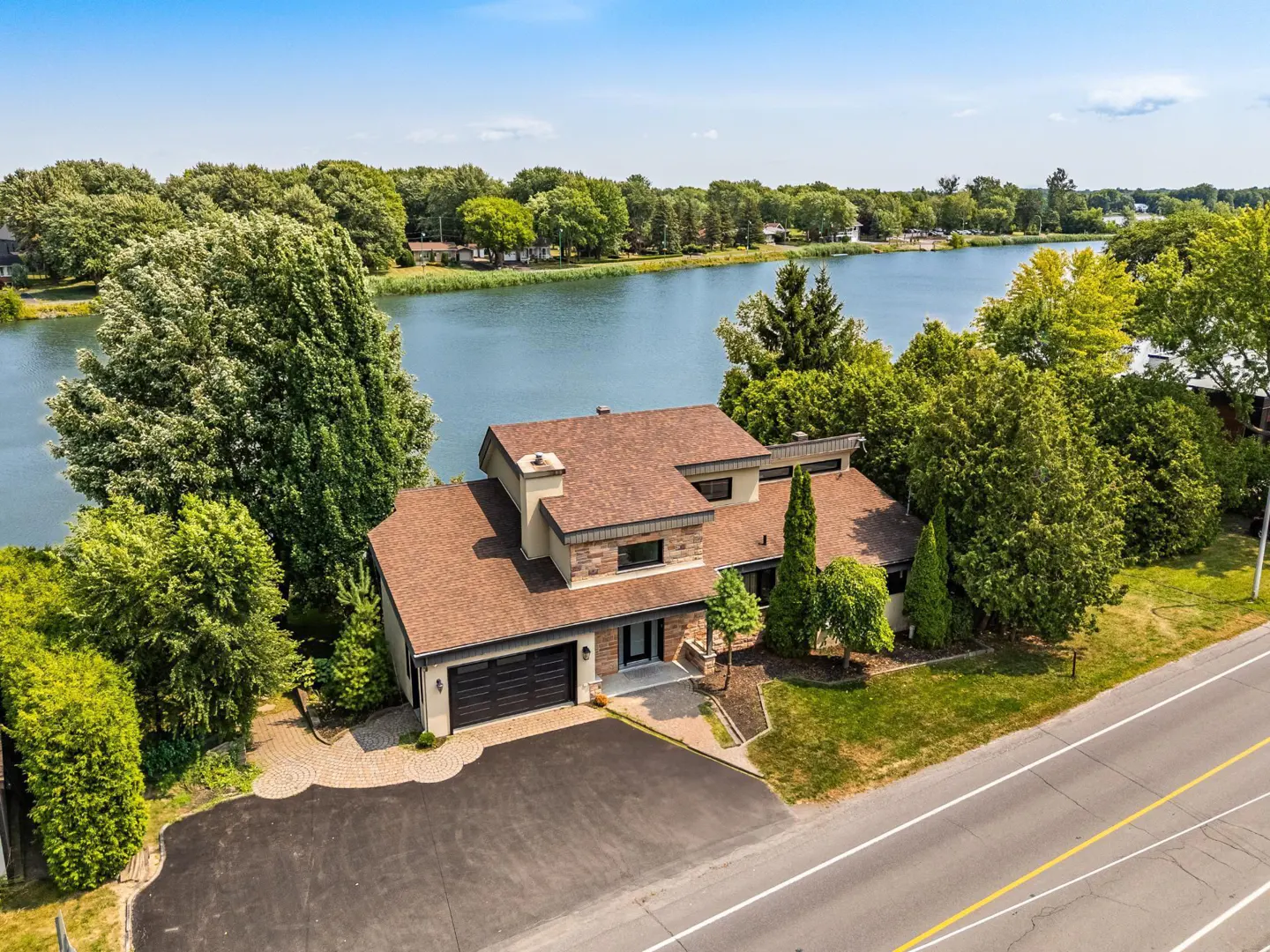 Aerial view of a modern two-story house with a brown roof, black garage doors, and a paved driveway, near a lake and a road.