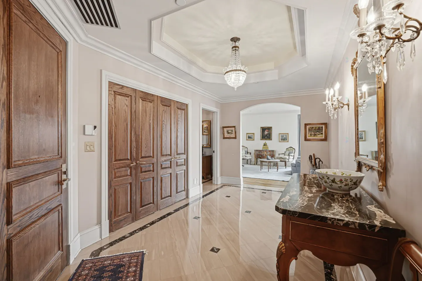 Elegant foyer with marble floors, wood doors, and a crystal chandelier. A table with a floral bowl sits near a gilded mirror.