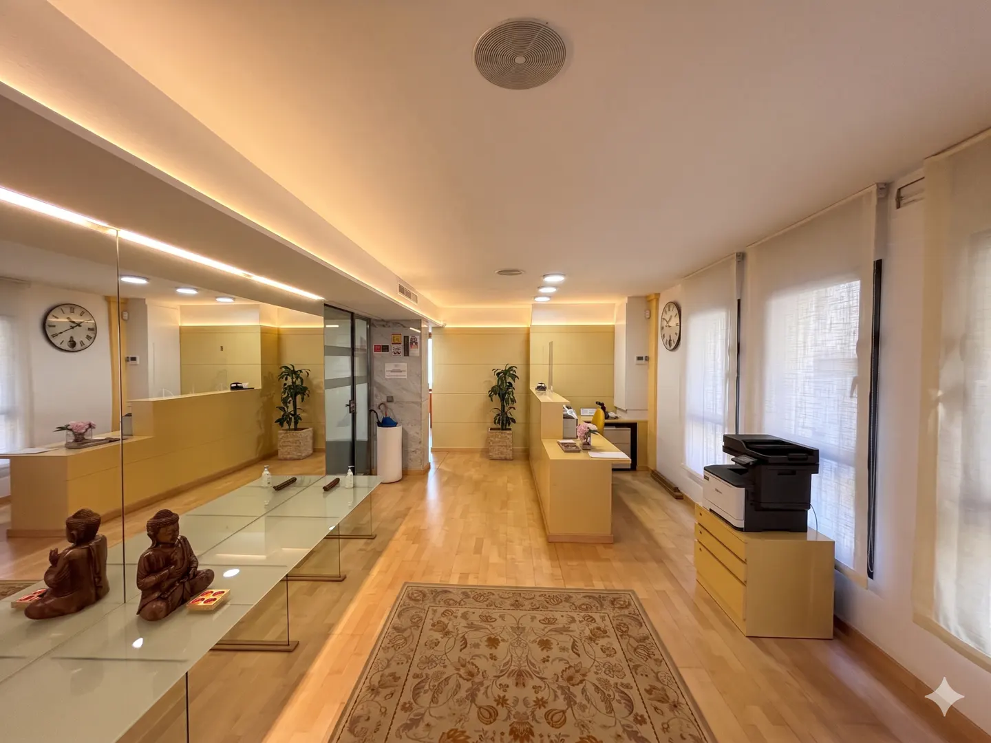 A bright office space with a long glass table, wooden floors, and a beige rug. Two wooden statues sit on the table.