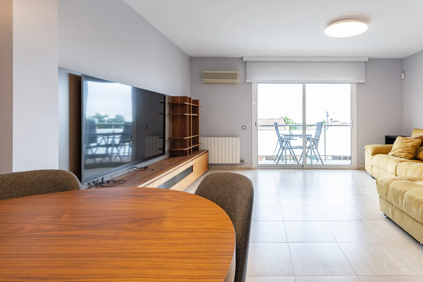 Living room with a wooden table and chairs, a large TV, and a balcony with outdoor furniture. The walls are gray, and the floor is tiled.