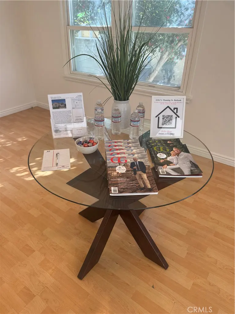A round glass table with a brown base holds magazines, water bottles, a plant, and real estate information in a bright room.