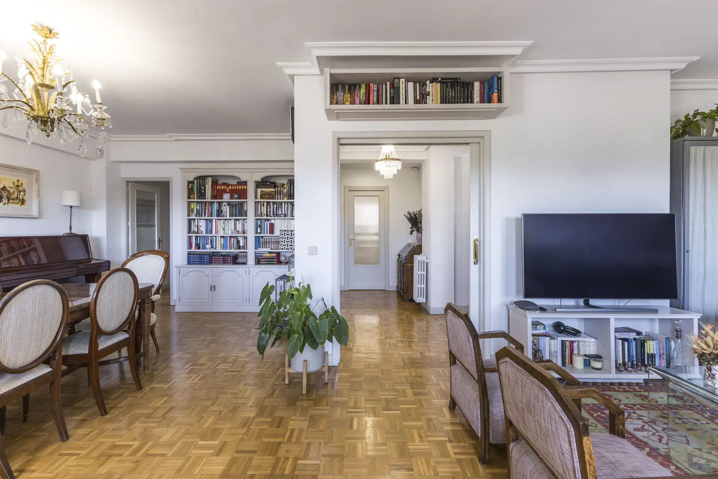 Bright living room with parquet floors, white walls, and bookshelves. A piano and dining table are on the left, and a TV and chairs are on the right.