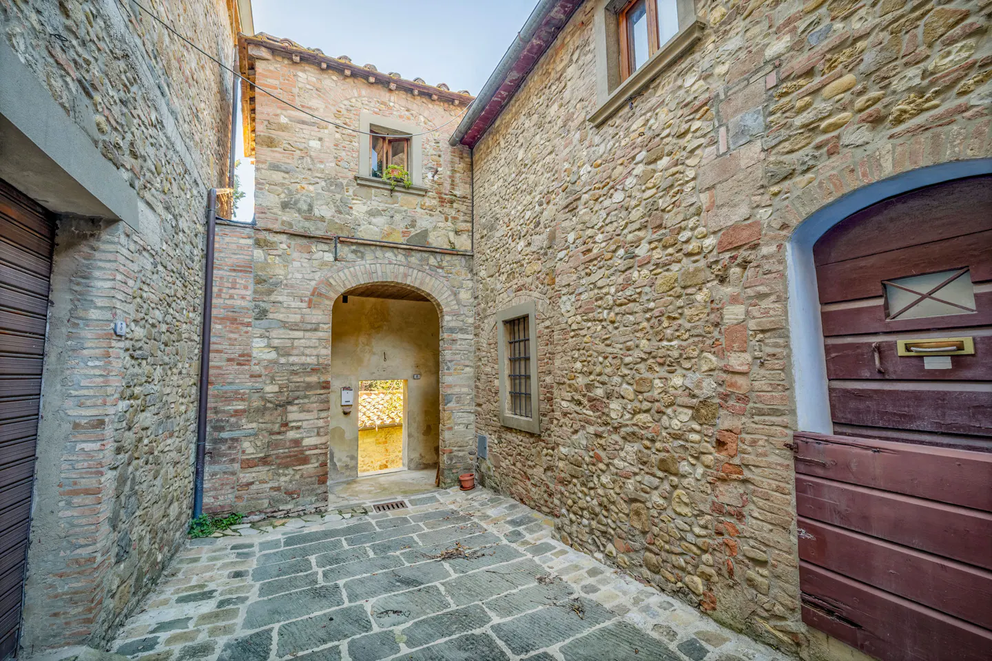 A narrow courtyard with rustic stone walls, a stone-paved floor, and a dark wooden door under a stone archway.