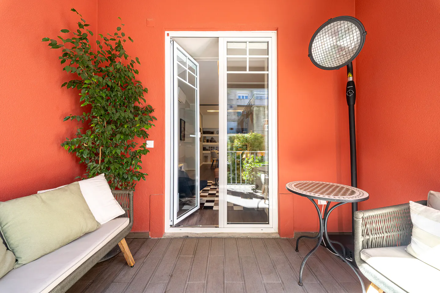 A balcony with orange walls, a white-framed door, a gray sofa with pillows, a small table, and a black heater.