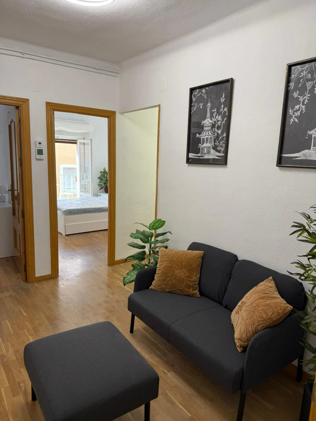 Living room with a gray sofa, ottoman, and wood floors. Two framed prints hang on the white wall. A bedroom is visible through a doorway.
