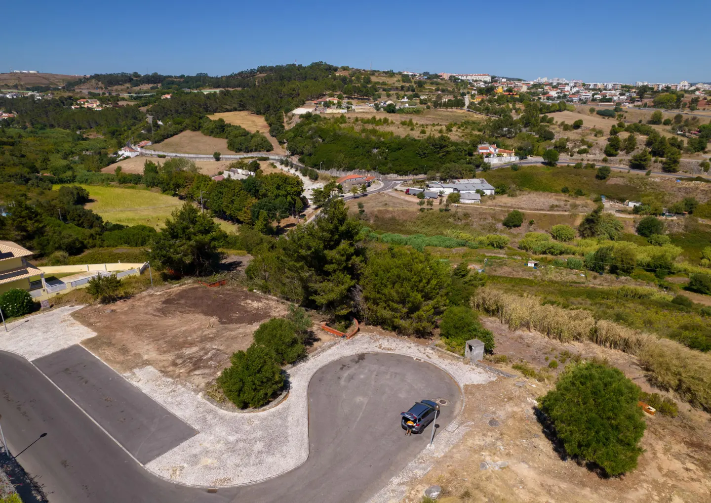 Aerial view of a paved cul-de-sac with a parked car, overlooking a rural landscape with hills, trees, and distant buildings.