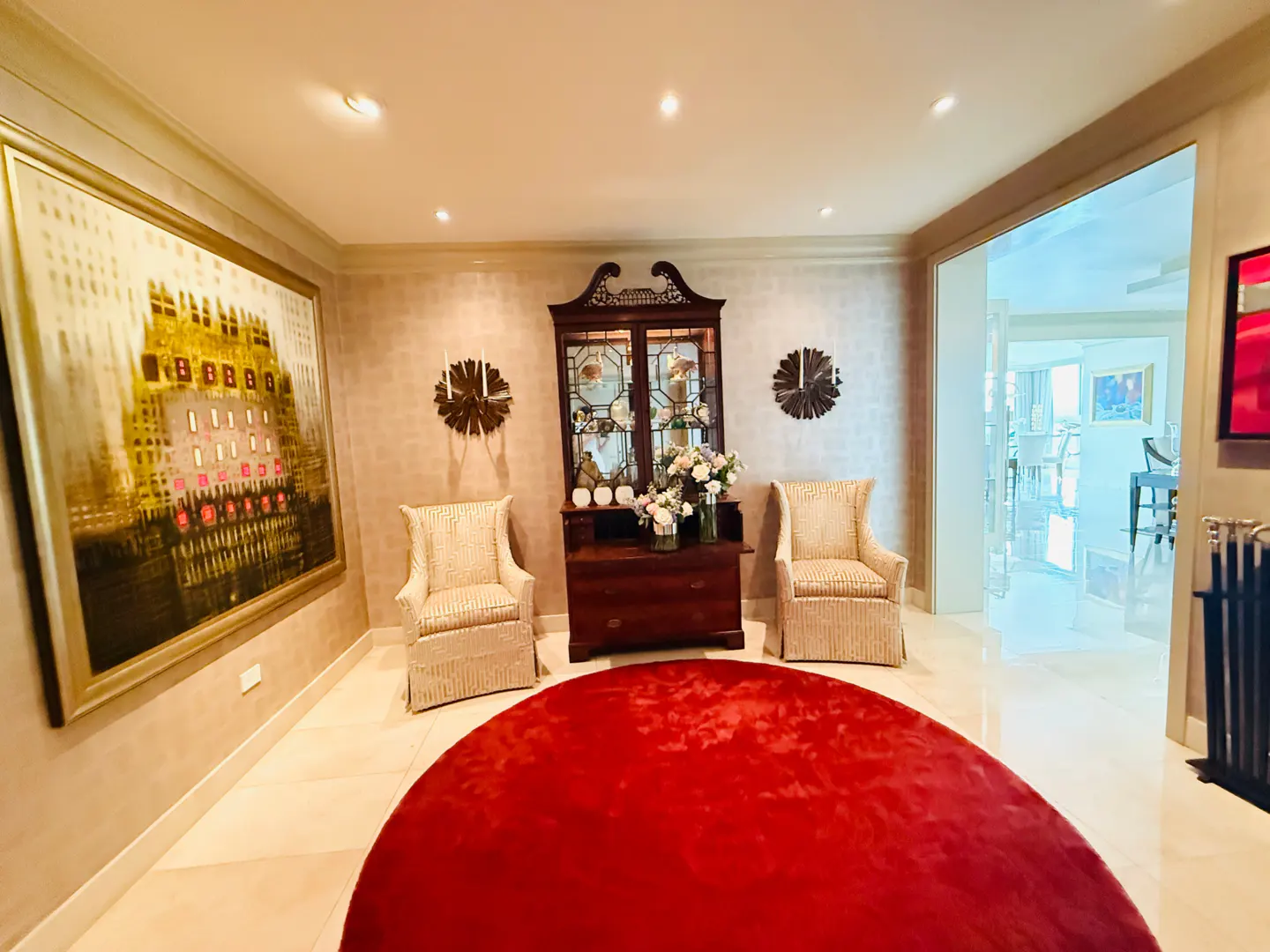 Elegant foyer with a red round rug, two chairs, and a dark wood cabinet. A large painting hangs on the left wall.
