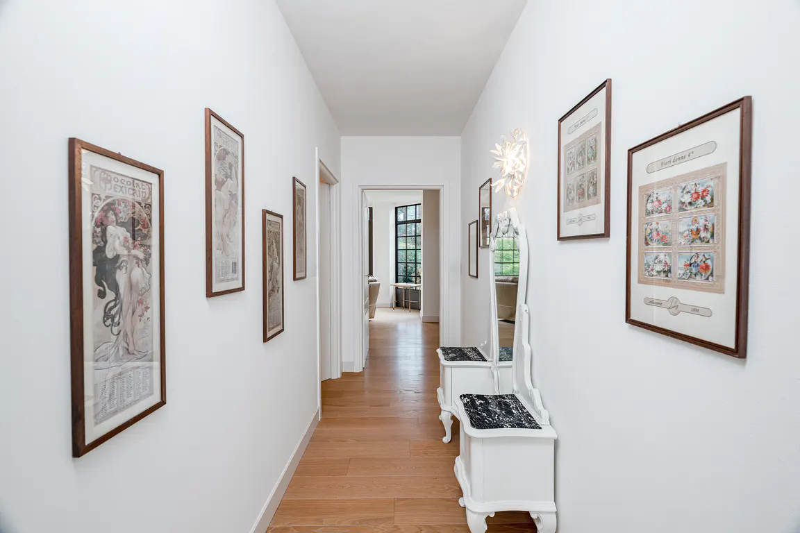 Bright hallway with white walls, wood floors, and framed art. A white vanity with a mirror sits on the right. A doorway leads to a living area.