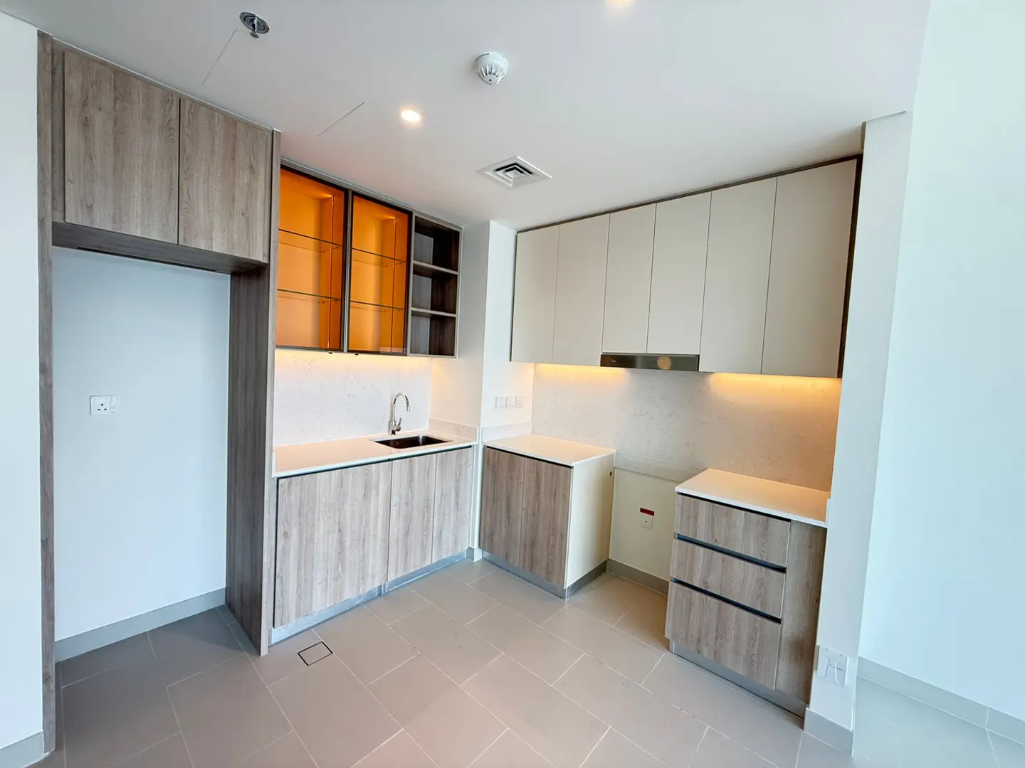 A modern kitchen with light wood cabinets, white countertops, and stainless steel sink. Orange glass cabinets are above the sink.