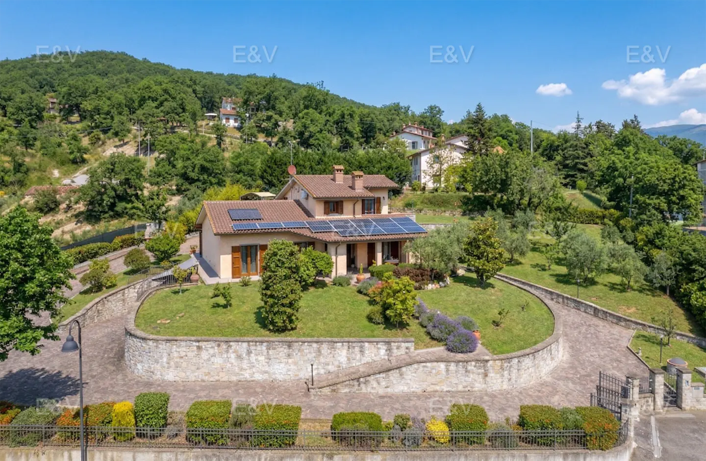 Exterior view of a beige house with a brown roof and solar panels, surrounded by green trees and a stone wall.