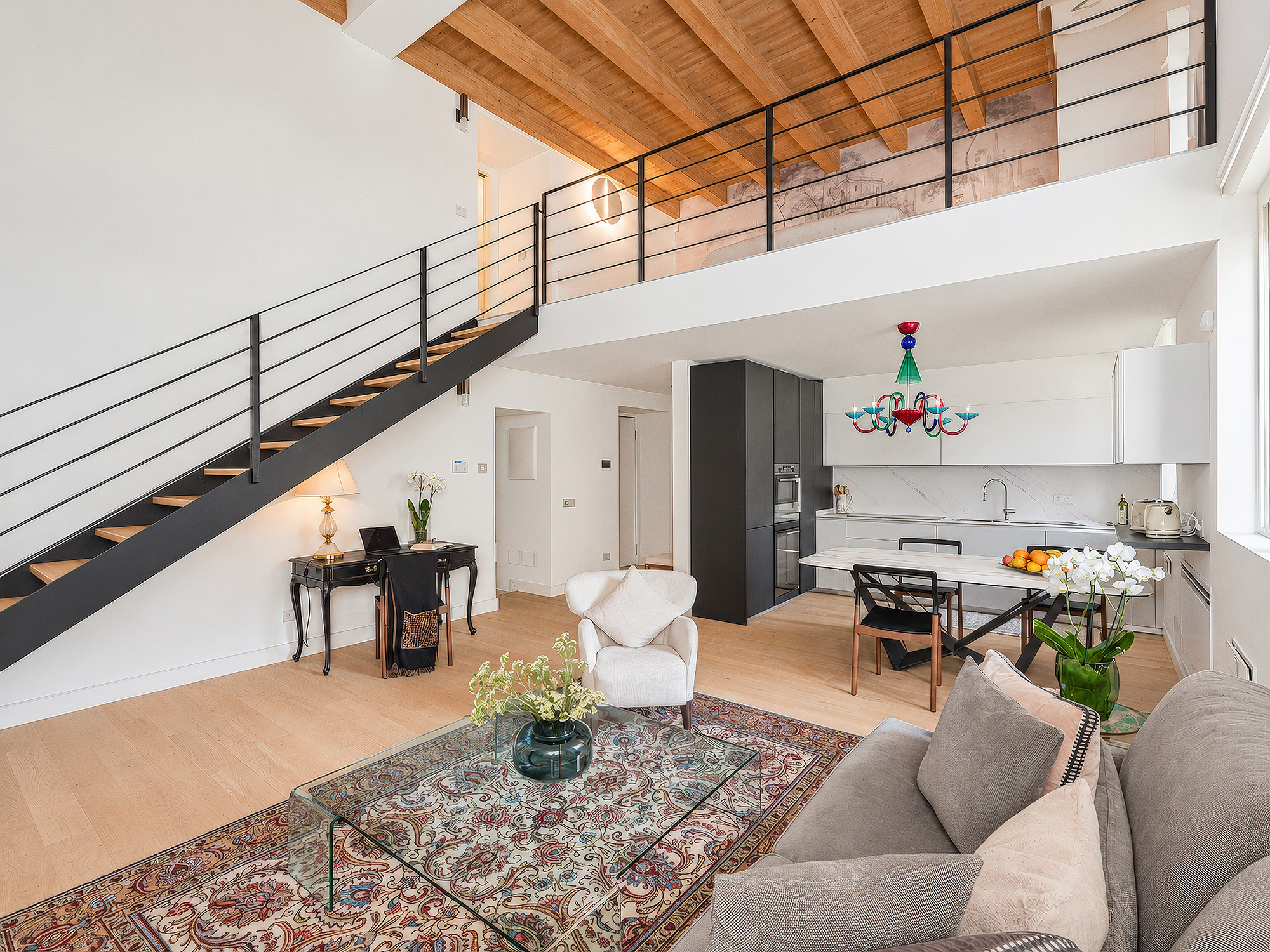 Modern open-plan loft with a black staircase, mezzanine, white kitchen, colorful chandelier, and a patterned area rug. [AI-generated text]