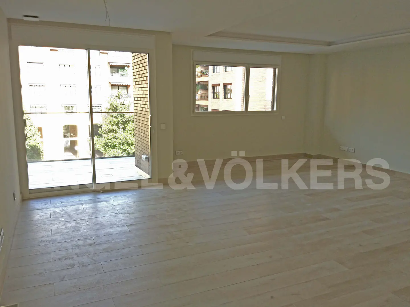 Unfurnished living room with light wood floors, beige walls, and large windows overlooking a brick building and trees.