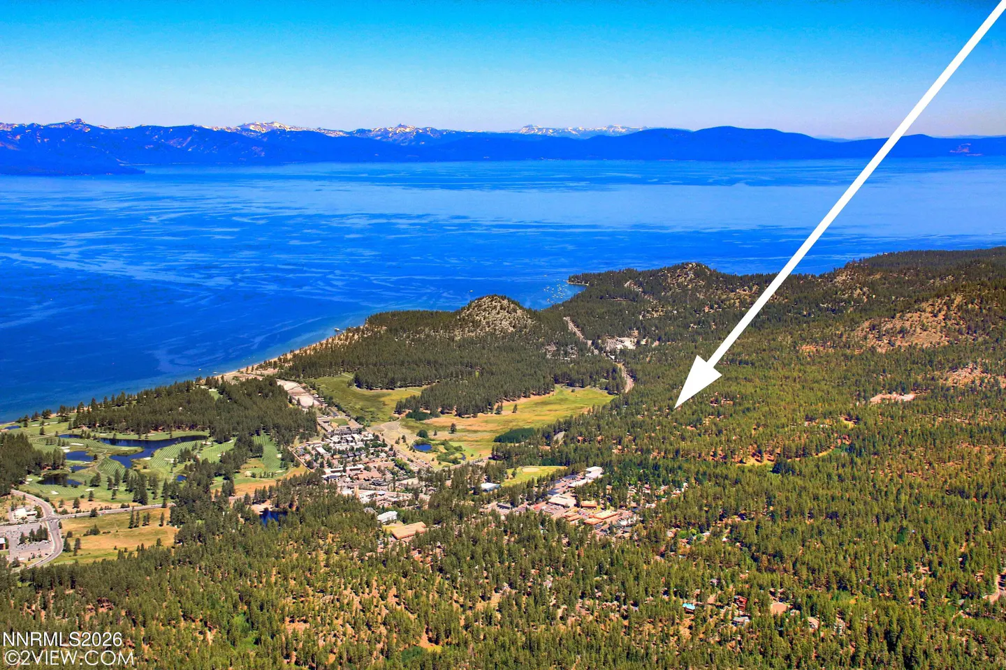 Aerial view of a town nestled in green trees near a blue lake and mountains under a clear blue sky.