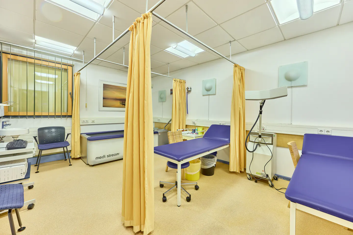 Bright medical exam room with blue exam tables, yellow privacy curtains, and medical equipment.