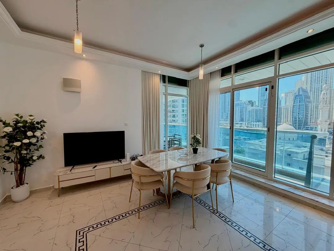Modern dining area with marble table, cream chairs, TV console, and floor-to-ceiling windows overlooking a city skyline.