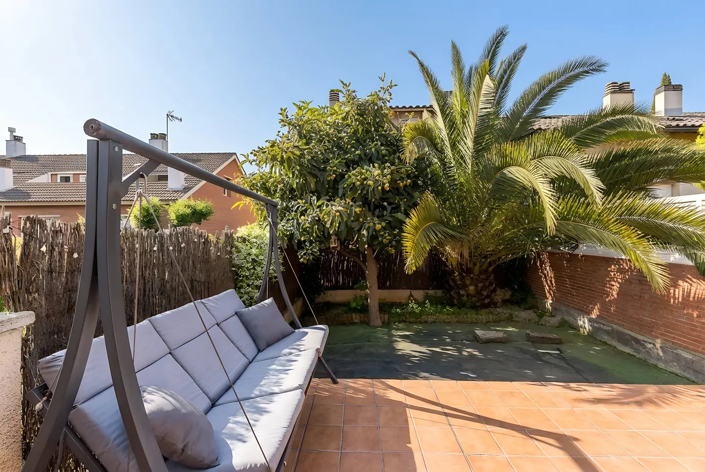 Outdoor patio with a gray swing sofa, brown tile flooring, and a green garden with trees and a brick wall.