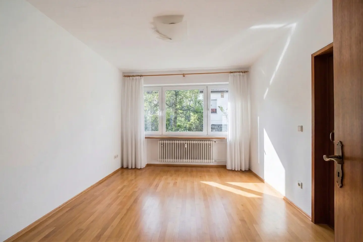 Bright, empty room with wood floors, white walls, and a window with white curtains. A radiator sits beneath the window. A partially open wood door is on the right.