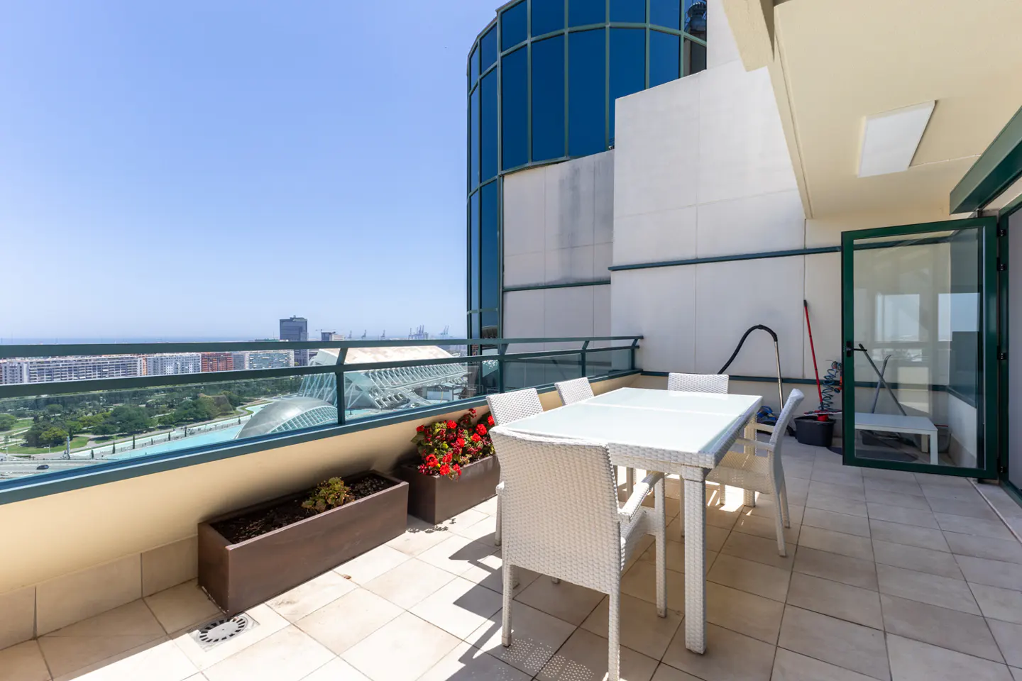 Modern balcony with white wicker dining set, potted red flowers, and a panoramic city view under a clear blue sky.