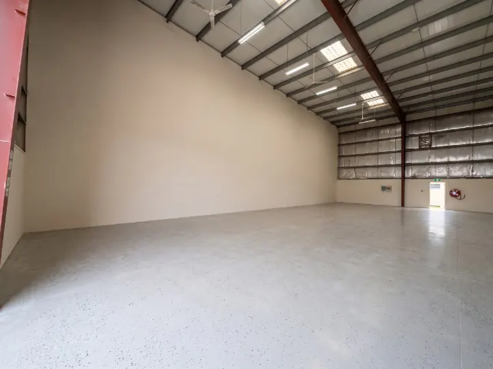 Empty warehouse interior with a concrete floor, white walls, and a high, metal roof with skylights and ceiling fans.