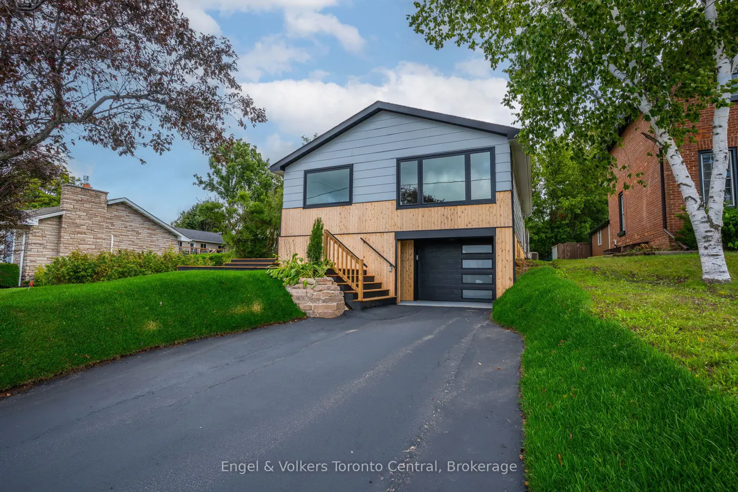 Two-story house with gray siding, wood paneling, black trim, and a black garage door with glass panels. A driveway leads to the garage.