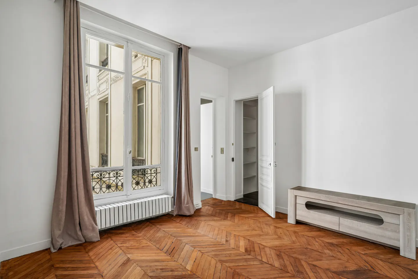 Bright, empty room with herringbone wood floors, white walls, and a large window with brown curtains. A white cabinet and open closet door are visible.