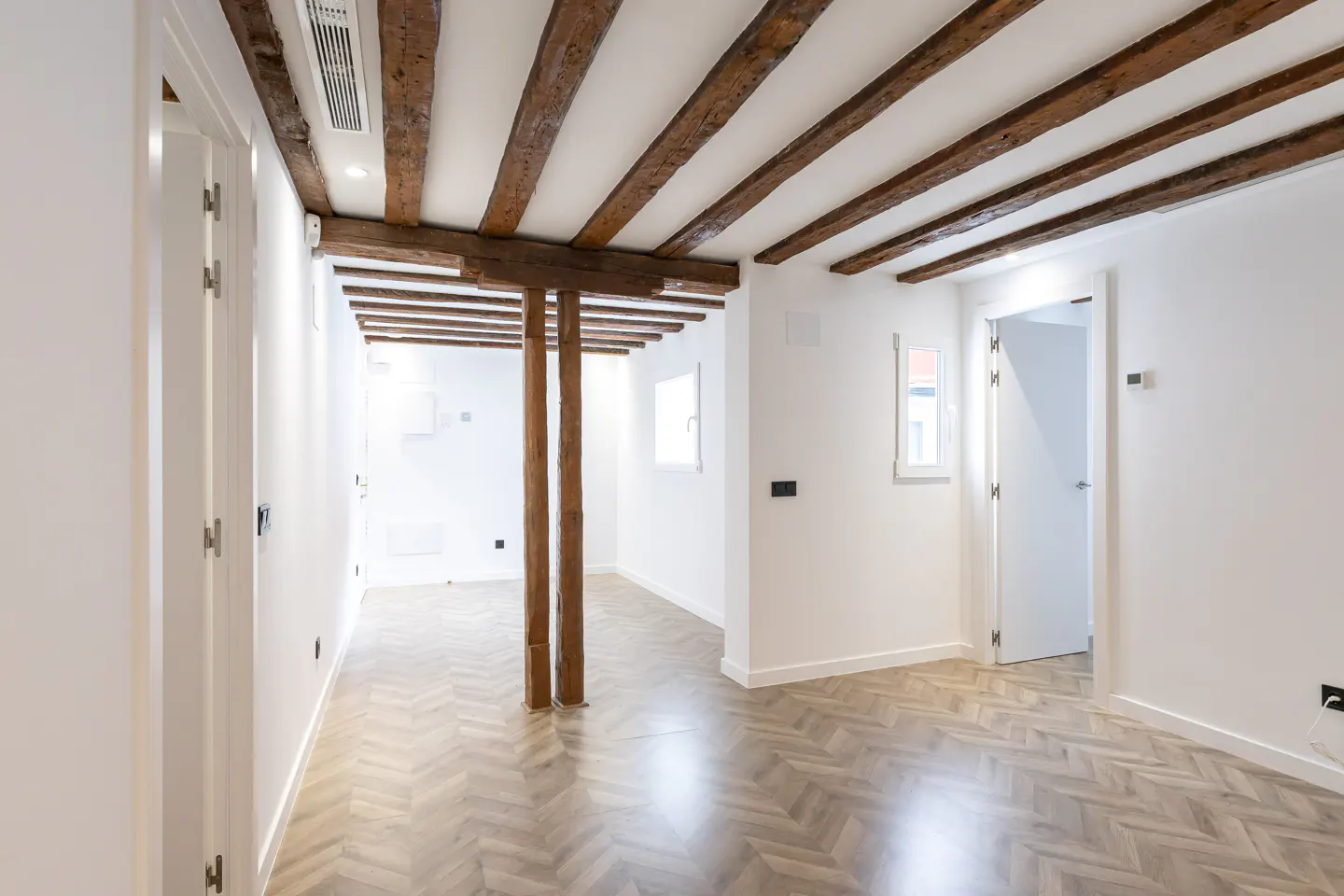 Bright, empty room with white walls, herringbone wood floors, and exposed dark wood beams and support posts.