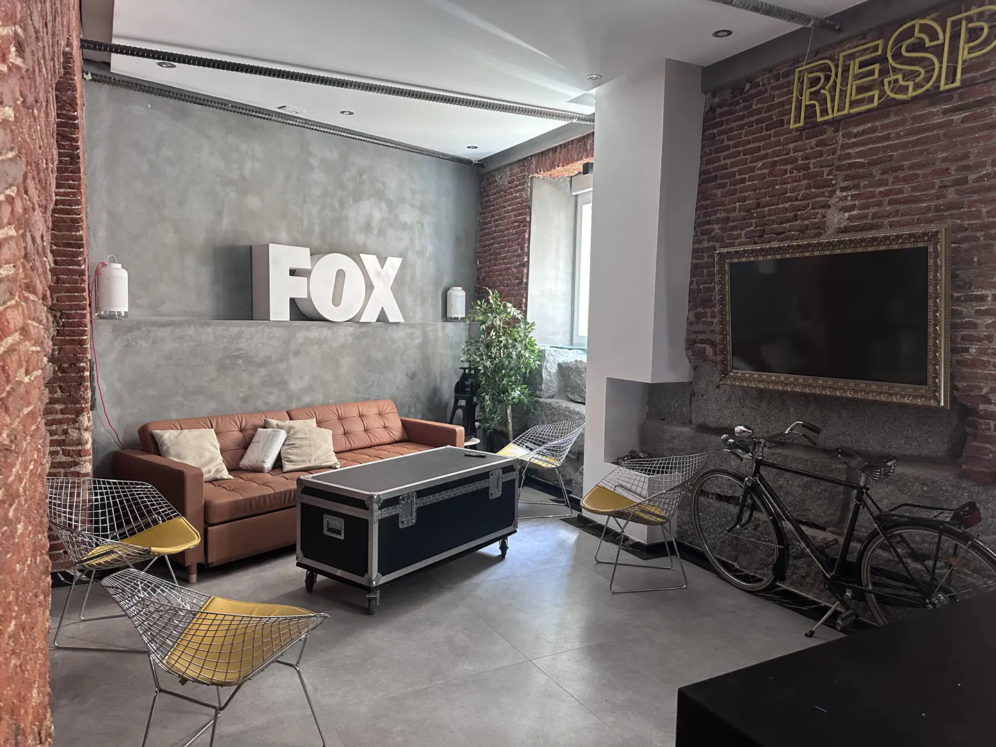 An industrial-style living room with exposed brick, a brown leather sofa, and metal chairs. A black trunk serves as a coffee table. A bicycle leans against the brick wall.
