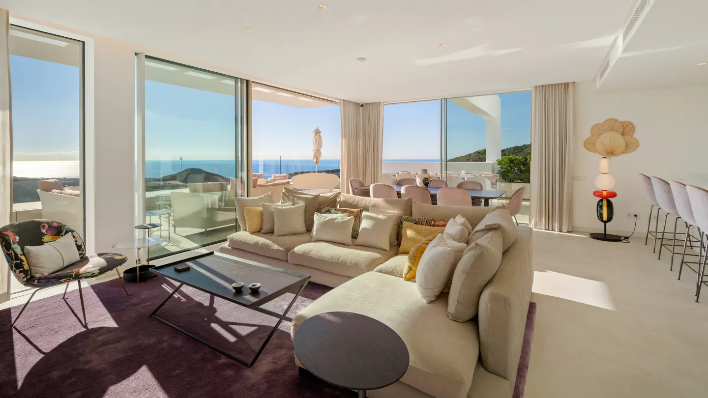 Bright living room with beige sectional sofa, purple rug, and ocean view through large windows. Dining area and bar stools visible.