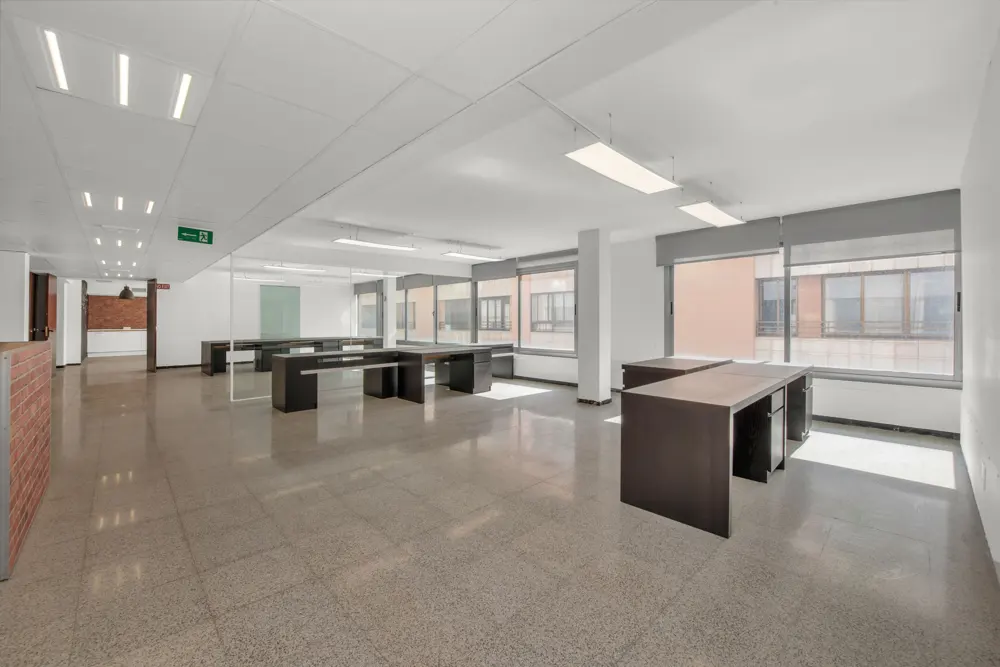 Bright, empty office space with dark desks, tiled floor, and large windows. An exit sign is visible in the background.