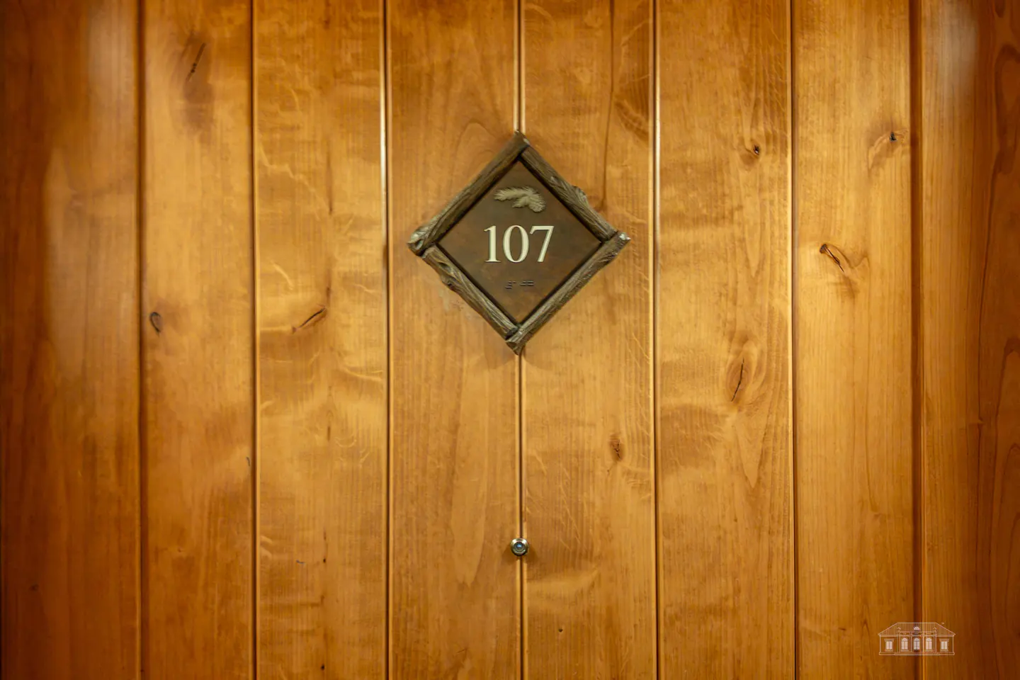 Wooden door with a diamond-shaped sign reading "107" in white numbers. The sign has a twig-like border and a small pinecone emblem.