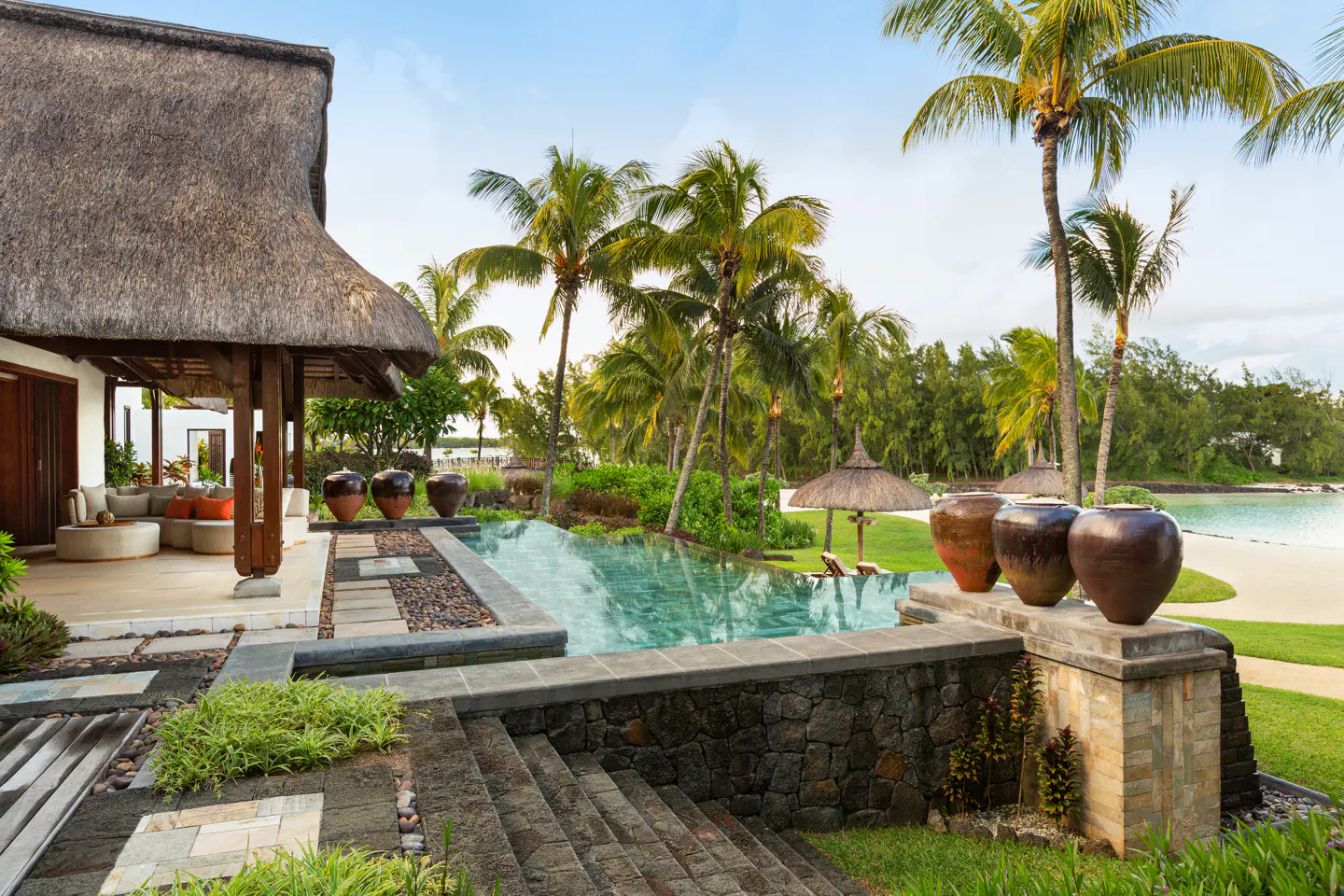 Luxury villa with thatched roof, infinity pool, and palm trees overlooking a white sand beach.