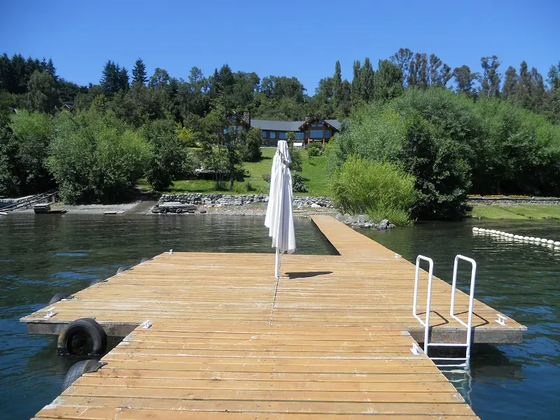 Wooden dock on a lake with a folded white umbrella and a ladder leading into the water. Trees and a house are in the background.