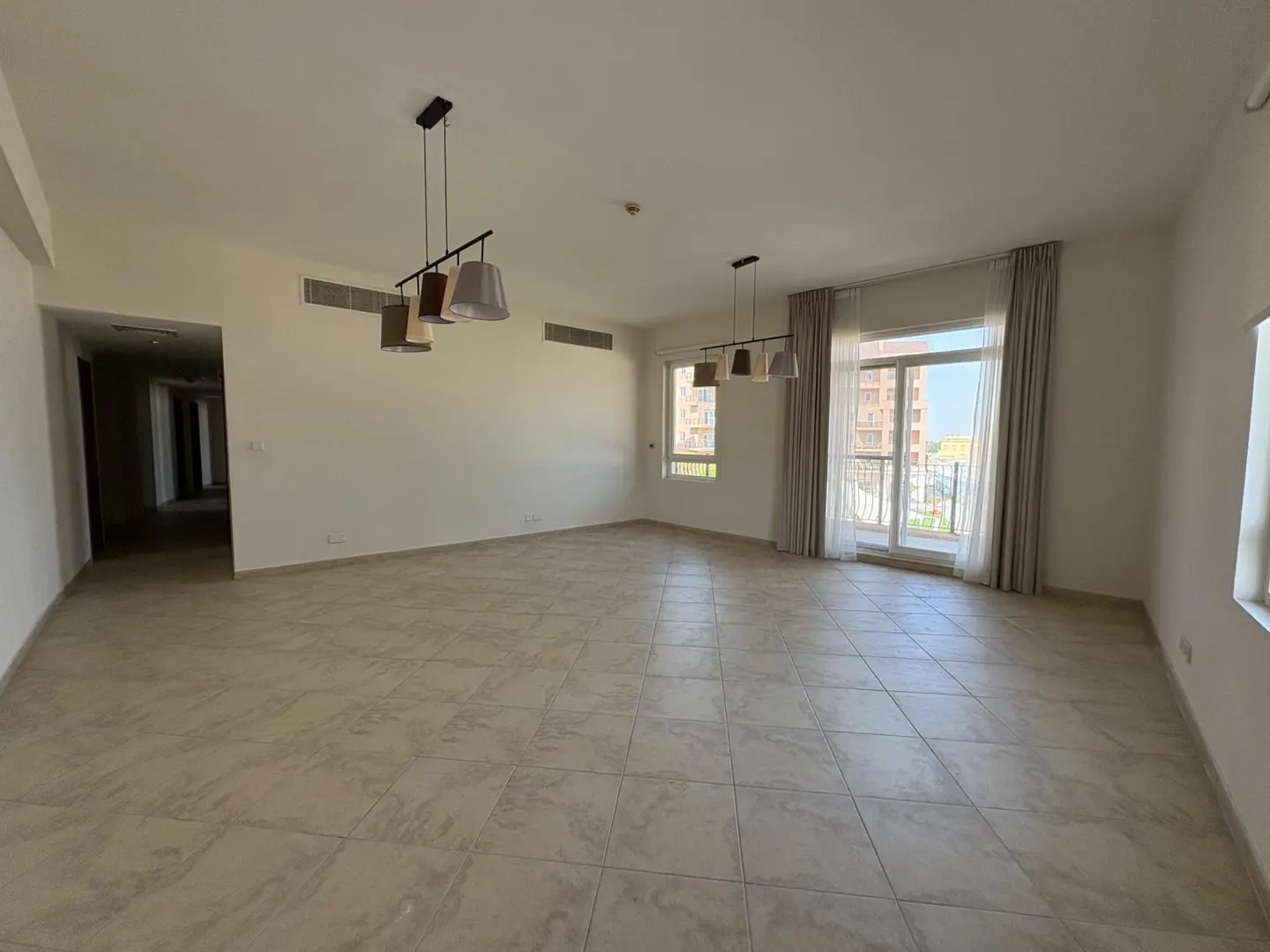 An empty, bright room with beige tile flooring, white walls, and two modern light fixtures. Windows with curtains offer a view of a building.