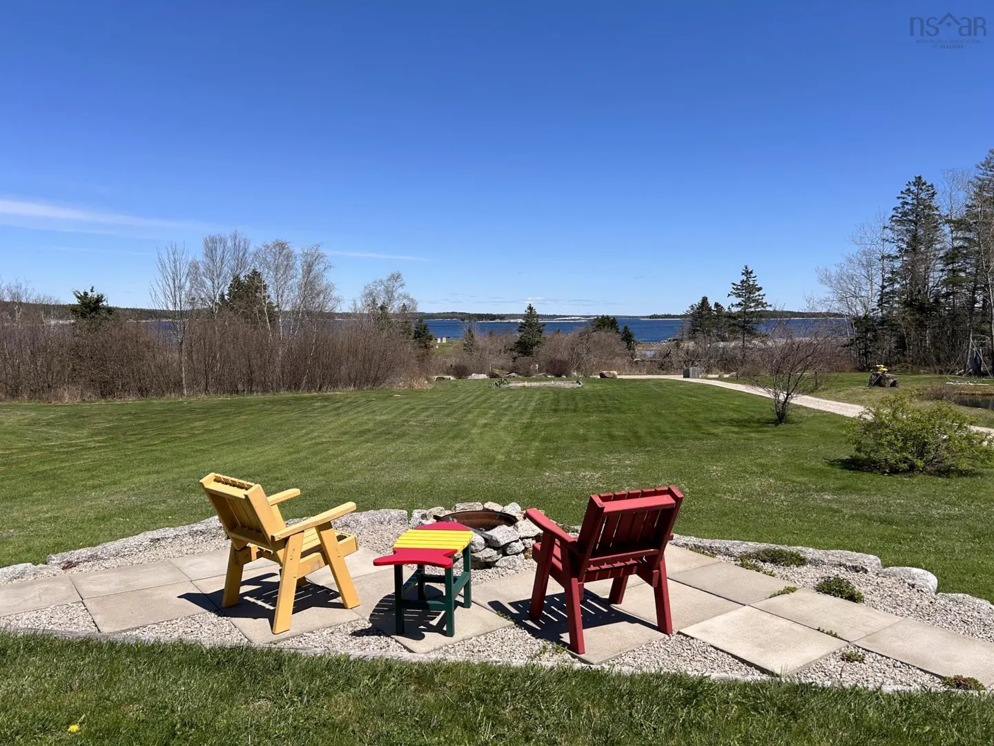 Outdoor patio with yellow and red Adirondack chairs, a colorful table, and a fire pit overlooking a green lawn and ocean view.