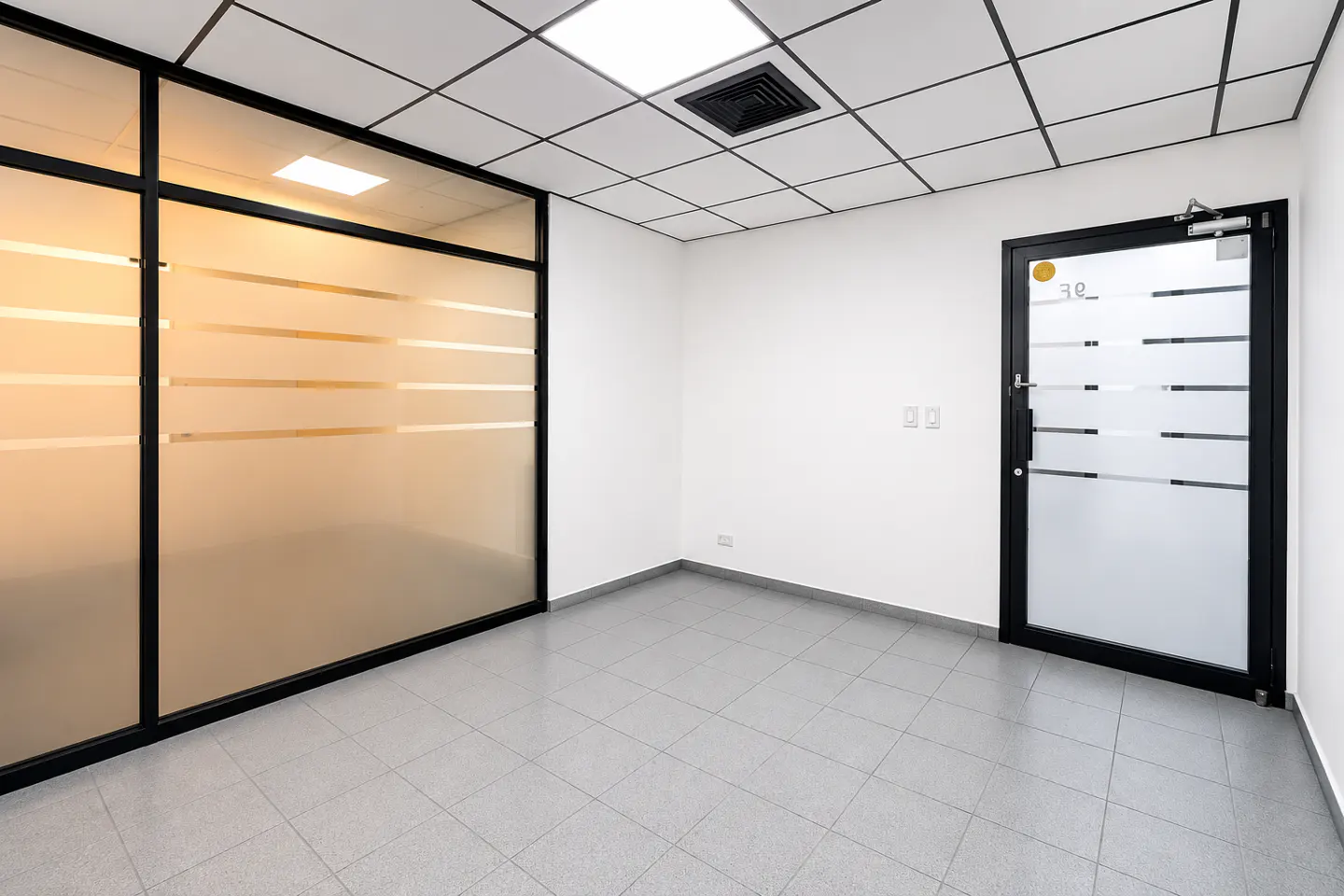 Empty modern office with white walls, grey tiled floor, drop ceiling, and a glass-walled room with a matching glass door.
