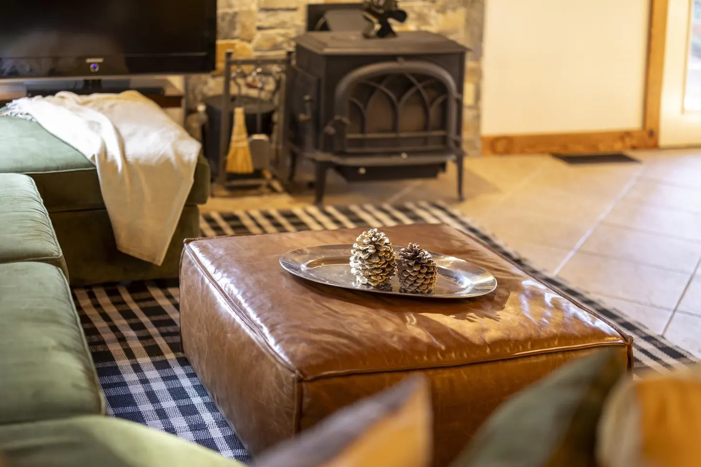 Living room with a brown leather ottoman, pine cones on a silver tray, green sofa, and black wood stove.