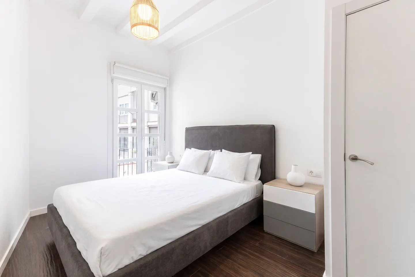 Bright bedroom with white walls, dark wood floors, and a gray bed. A window and a white door are visible.