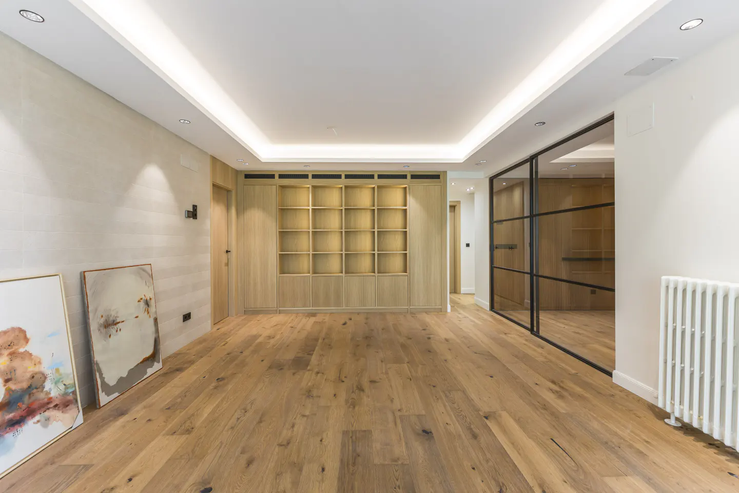 Bright, empty room with wood floors, built-in shelves, and two paintings leaning against the wall. Black framed glass doors are on the right.