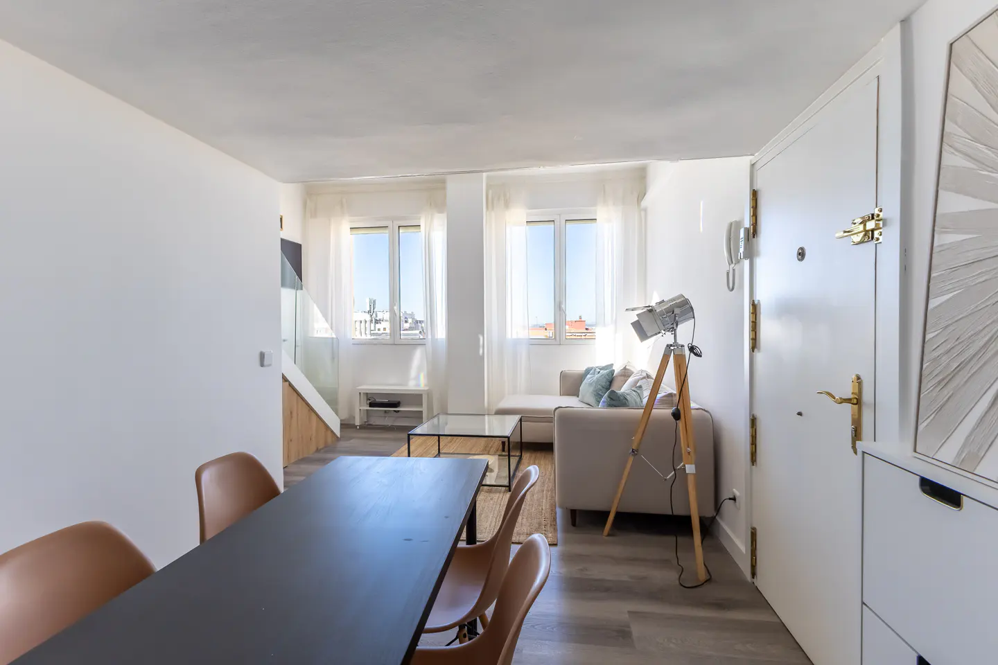 Bright apartment interior with a black dining table, tan chairs, a beige sofa, and a vintage spotlight on a wooden tripod.